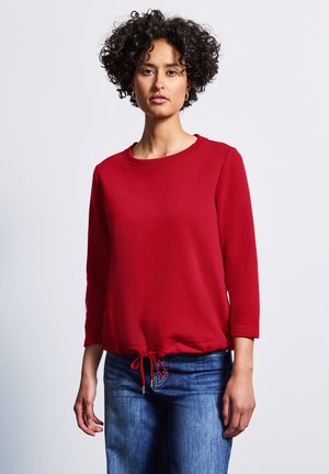 Pullover rosso con scollo a giro, maniche a tre quarti e orlo con coulisse. Tessuto morbido con vestibilità ampia. Indossato con jeans blu.