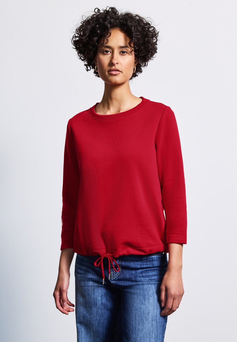 Röd sweatshirt med rund halsringning, trekvartslånga ärmar och dragsko i nederkant. Mjuk textur med en lös passform. Bärs med blå jeans.