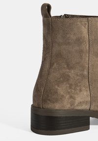 Bottine en daim marron avec une finition texturée, bout arrondi et un talon en caoutchouc noir bas. Fermeture éclair sur le côté et tirant à l'arrière.