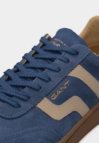Μπλε σουέτ αθλητικό παπούτσι με μπεζ δερμάτινες λεπτομέρειες, μπλε κορδόνια και χρυσό λογότυπο "GANT" στο πλάι πάνω από καφέ σόλα.