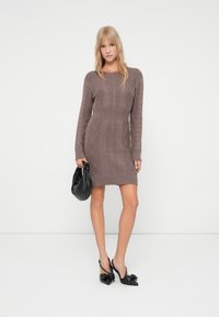 Rochie tricotată în nuanțe de maro estompat, cu un model de tricotaj cu cabluri, mâneci lungi, talie ajustată și un tiv scurt. Purtați cu accesorii negre.