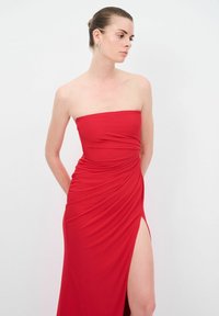 Červené maxi šaty bez ramínek s nabíranou texturou, vysokým rozparkem na jedné straně a hladkým, elastickým materiálem. Elegantně se přizpůsobují postavě.