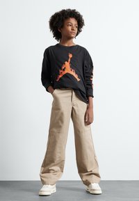 Camisa negra de manga larga con gráfico y texto naranja en las mangas, combinada con pantalones cargo beige de pierna ancha y zapatillas blancas.