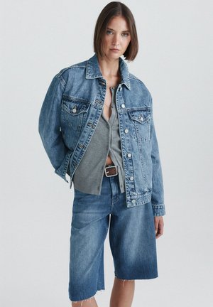 Jeune femme portant une veste en jean bleue oversize et un short long en jean assorti par-dessus un haut gris boutonné, debout devant un fond uni.