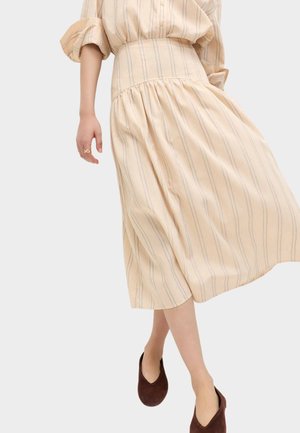 Jupe midi beige à rayures verticales associée à une chemise assortie aux manches retroussées et des chaussures plates marron, portée par une femme en train de marcher.