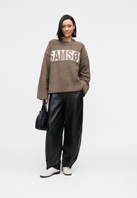 Camisa castanha de malha com texto branco contrastante, combinada com calças largas de couro preto e sapatos deslizantes de cor clara. Pequena mala preta.