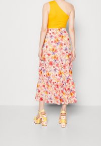 Frau steht, trägt ein gelbes Ein-Schulter-Oberteil, einen geblümten Midirock mit rosa, lila und orangefarbenen Blumen sowie gelbe, blumige High-Heels.