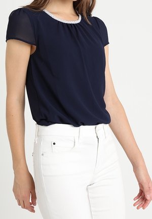 Femme portant un chemisier bleu marine à manches courtes avec un liseré blanc et un pantalon blanc taille haute, debout devant un fond uni.