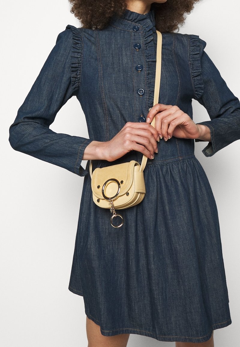 Vestito in denim con dettagli arricciati sulle spalle, bottoni blu e vita arricciata. Piccola borsa a tracolla gialla con chiusura a cerchio.