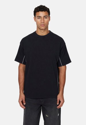 Schwarzes T-Shirt mit kurzen Ärmeln, lockerer Passform, Rundhals und sichtbaren weißen Nähten an den Seitennähten. Kombiniert mit abgewetzten dunkelblauen Jeans.