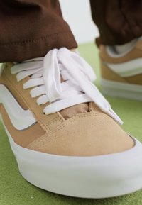 Tan suede sneaker with white laces and a white side stripe. Smooth texture, rounded toe, and contrasting rubber sole.