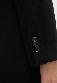 Manica di cappotto in lana nera con tre bottoni neri lucidi, tessuto testurizzato e dettagli di cucitura discreti. Finitura liscia con un design aderente.