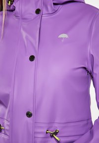 Fioletowa wodoodporna kurtka wykonana z gładkiego materiału, z czarnymi guzikami, srebrnym logo parasola oraz regulowanymi troczkami z żółtymi akcentami.