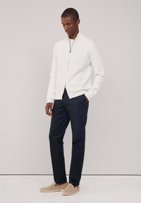 Cardigan bianco in maglia a coste con chiusura a zip, indossato sopra una maglietta bianca, abbinato a pantaloni blu navy e scarpe slip-on beige.
