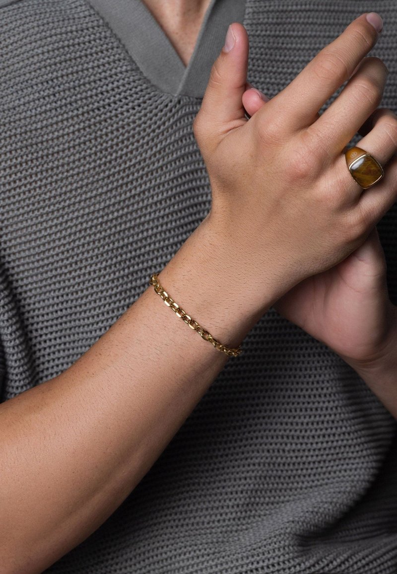 Guld kædearmbånd på et håndled, der har sammenkoblede led. Den strukturerede grå beklædning tilføjer kontrast til det glatte metaldesign.