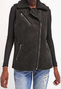 Gilet senza maniche in shearling nero con zip asimmetrica e tasche con zip indossato sopra una maglia nera a maniche lunghe e jeans strappati blu.