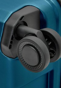 Hartschalentrolley in Blau mit strukturierter Oberfläche. Verfügt über einen schwarzen Radmechanismus mit einem glatten, runden Rad und gerilltem Rand.