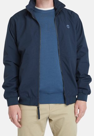 Mann trägt eine marineblaue Jacke mit Reißverschluss über einem blauen Pullover und beigen Hosen, steht mit entspannten Händen an den Seiten.