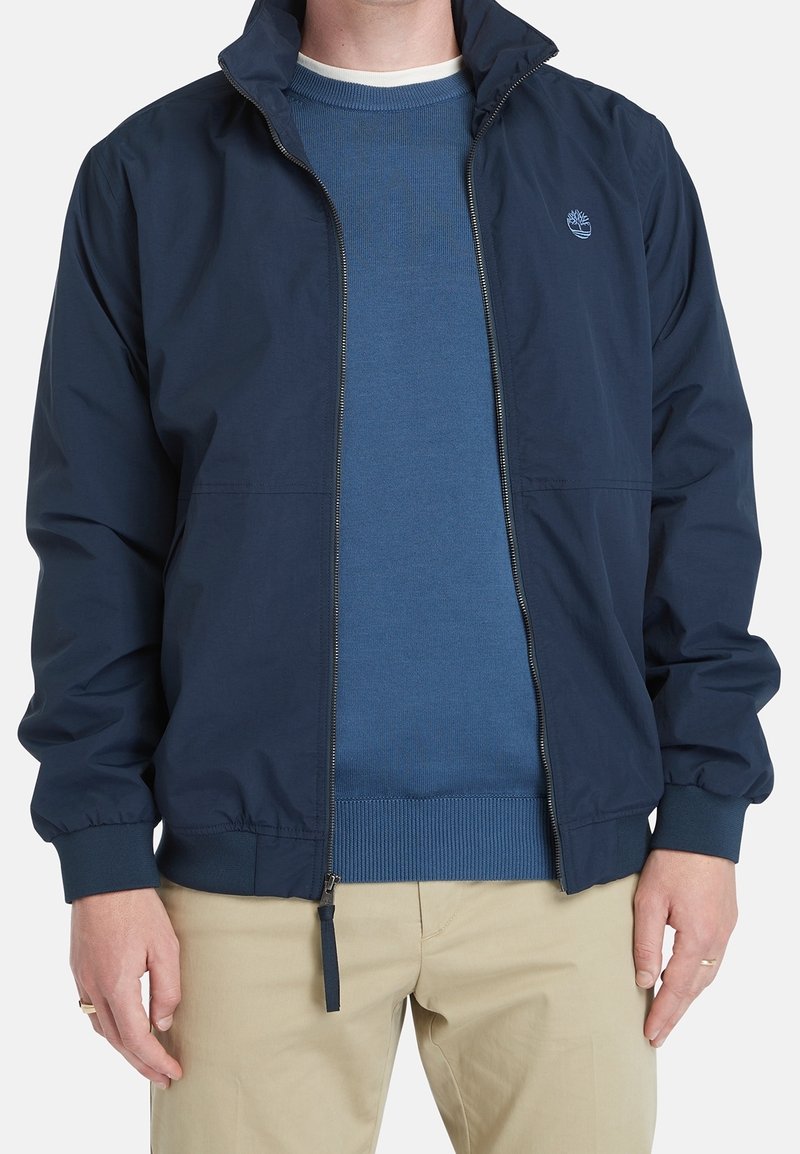 Homme portant une veste zippée bleu marine par-dessus un pull bleu et un pantalon beige, debout les mains détendues le long du corps.