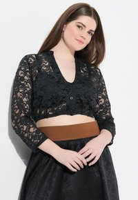 Schwarzes Spitze-Crop-Top mit langen Ärmeln, V-Ausschnitt und Knopfverschluss. Kombiniert mit einem hochtaillierten Rock mit einem braunen elastischen Bund.