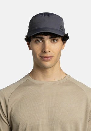 Joven con cabello rizado oscuro que lleva una gorra gris y una camiseta beige de manga larga con textura, mirando al frente con expresión neutral.