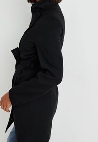 Zwarte blazer met een getailleerde silhoutte, hoge kraag en een vastgebonden taille-detail. Beschikt over lange mouwen en een zachte textuur, geschikt voor het laagjes dragen.