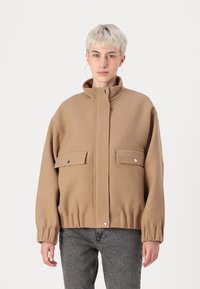 Hnědá, oversized bunda vyrobená z texturované tkaniny, s vysokým límcem, dvěma předními kapsami se zapínáním na druky a elastickým lemem.