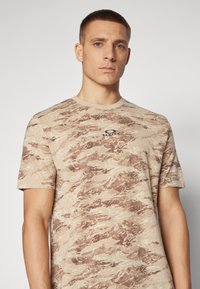 Beige lyhythihainen t-paita, jossa on teksturoitu ruskea ja krem camouflage-kuvio, ja pienellä mustalla "Oakley"-logolla rinnassa.