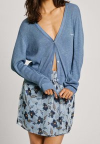 Cardigan a coste azzurro chiaro con scollatura a V profonda e maniche lunghe, abbinato a una gonna mini con motivo floreale che presenta fiori e foglie blu.