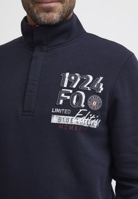 Pullover blu navy con colletto alto, caratterizzato da testo bianco e rosso: "1924 FQ Edizione Limitata" e un patch rotondo con logo. Texture del tessuto morbida.