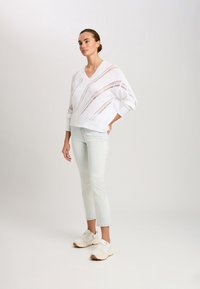 Weißer Strickpullover mit V-Ausschnitt, der offene Muster aufweist. Kombiniert mit hellblauen Slim-Jeans und weißen Turnschuhen, steht das Modell im Profil.