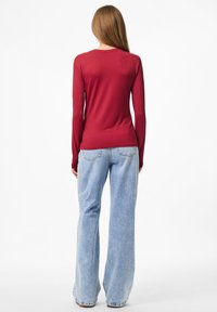 Vrouw met lang bruin haar, gekleed in een strakke rode top met lange mouwen en lichtblauwe wijde jeans, staand met haar rug naar de camera tegen een witte achtergrond.