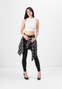 Witte fitted crop top, zwarte skinny jeans en een zwart-wit geruite blouse om de taille geknoopt. Zwarte hoge hakken.