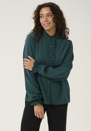 Dark green blouse made of soft fabric with a ruffled collar, button-up front, and gathered sleeves. Paired with black pants.