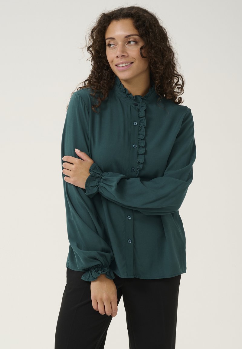 Blusa verde scuro realizzata in tessuto morbido con colletto arricciato, chiusura a bottoni e maniche gather. Abbinata a pantaloni neri.