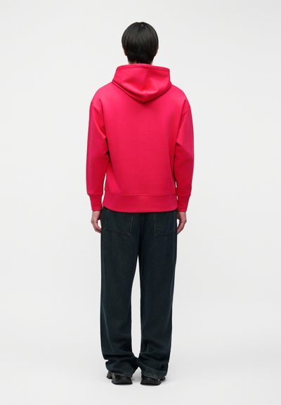 Άτομο με κοντά μαύρα μαλλιά που φοράει φωτεινή κόκκινη κουκούλα και φαρδιά σκούρα παντελόνια, στέκεται με την πλάτη γυρισμένη σε απλό φωτεινό φόντο.