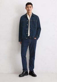 Man in een donkerblauwe spijkerjacket en jeans met een wit shirt en zwarte schoenen, staande tegen een witte gestructureerde muur.