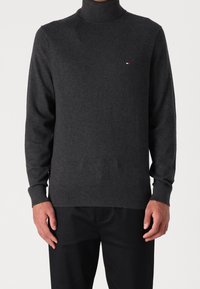 Grauer Rollkragenpullover aus weichem Material, mit langen Ärmeln und einem kleinen Logo in Rot, Weiß und Blau auf der Brust.