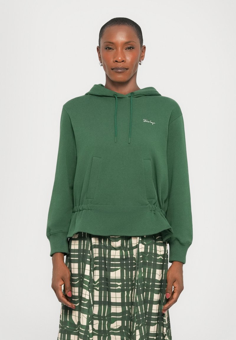 Felpa verde con cappuccio e vestibilità rilassata, dotata di due tasche anteriori e dettagli arricciati sottili all'orlo. Indossata sopra una gonna a scacchi.
