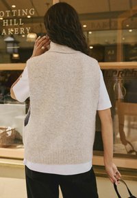 Gilet lavorato a maglia color beige chiaro con colletto alto, orlo a coste e tasche laterali, indossato sopra una camicia bianca e abbinato a pantaloni neri.