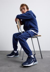 Mörkblå hoodie och joggers, mjukt tyg, med vit Nike-logotyp. Svarta och mörkblå sneakers med nätöverdela och genomskinlig sula.