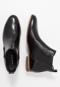 Botas de cuero negro hasta el tobillo con una superficie lisa, paneles laterales elásticos y un tacón de madera bajo. Nombre de la marca "ANNA FIELD" visible en el interior.