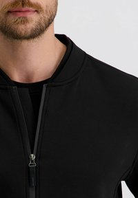 Schwarze Sportjacke mit Reißverschluss und hohem Kragen. Glattes Material mit dezenter Glanzoberfläche. Verfügt über kontrastierende Nähte und ein Logotag.
