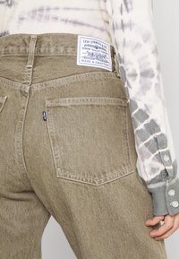 Vaqueros de mezclilla verde oliva con cintura media y cuatro bolsillos. Presentan una etiqueta azul de Levi's en la parte trasera, un acabado sutilmente texturizado y detalles de costura.