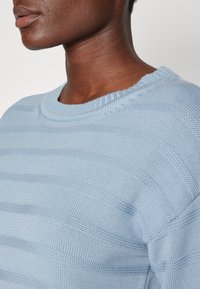 Maglione di colore azzurro chiaro realizzato a maglia, con motivi testurizzati orizzontali, colletto a girocollo a costine e un tessuto morbido e traspirante.
