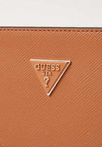 Ruskea tekstuurinen materiaali, jossa on kolmiomainen logo-tagi, jossa lukee "GUESS" ja "USA" kultaisella kirjasimella, jota korostaa kultainen kysymysmerkki-symboli.