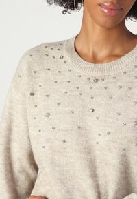 Maglione beige realizzato in morbido tessuto a maglia, caratterizzato da decorazioni metalliche sparse sulla parte anteriore e un collo a girocollo rigato.