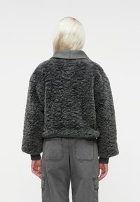 Person med langt, lyst hår iført en mørkegrå tekstureret fleecejakke og matchende cargo-bukser, vist fra ryggen mod en ensfarvet baggrund.