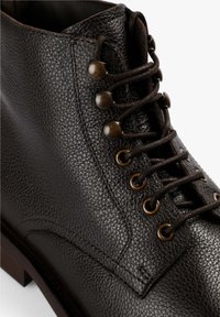 Bota de tobillo de cuero marrón con una superficie texturada, ojales de latón y cordones marrones. Presenta una suela de madera sólida y un contraste en la costura.