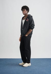 Μαύρο φούτερ με φερμουάρ και κόκκινο-λευκό λογότυπο, φορεμένο πάνω από λευκή αμάνικη μπλούζα, συνδυασμένο με μαύρο παντελόνι και λευκά αθλητικά παπούτσια.